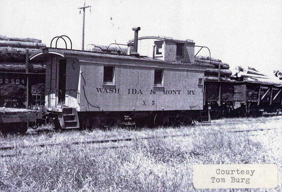 View of a WI&M railroad car. Behind the car, stacks of logs can be seen being hauled in more railroad cars.