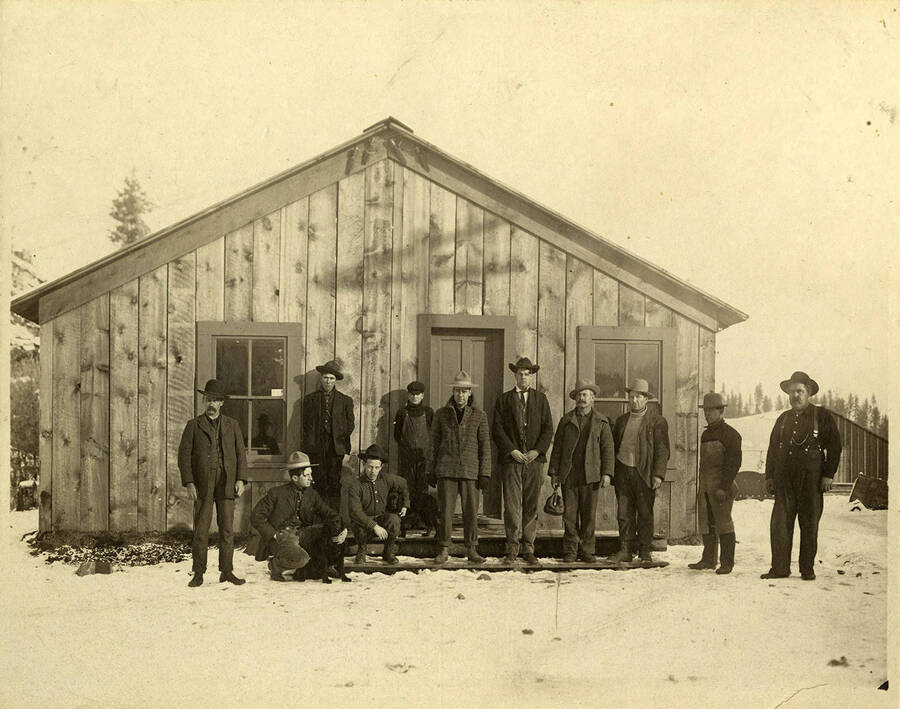 Eleven men outside a wood building.