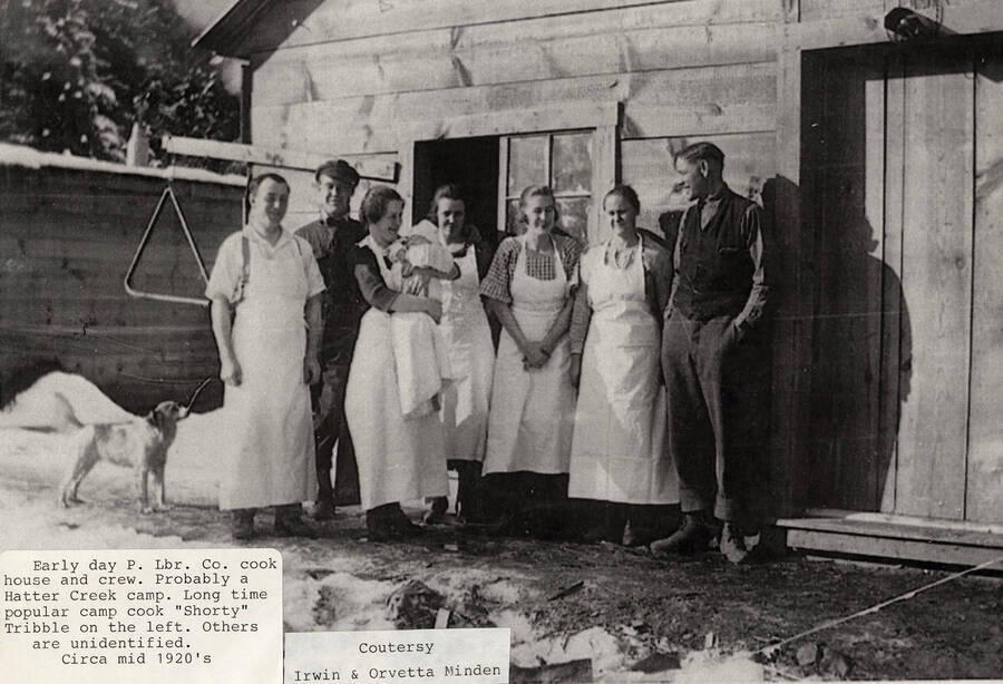 The cooking crew standing in front of a cookhouse at a Hatter Creek camp. A dog can be seen standing under a large triangle.