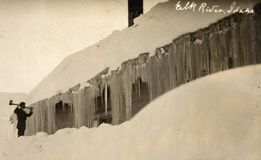 A photograph of a man using his axe to chop down massive icicles from a building in Elk River, Idaho.