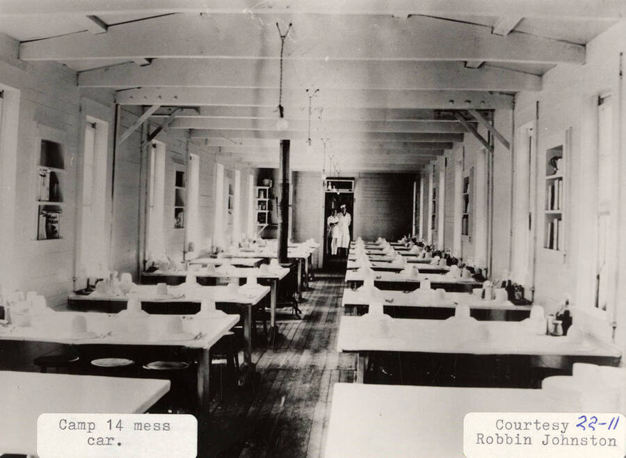 View of the mess car at PLC camp 14. The tables can be seen set with dishes and two cooks can be seen standing at the end of the car.