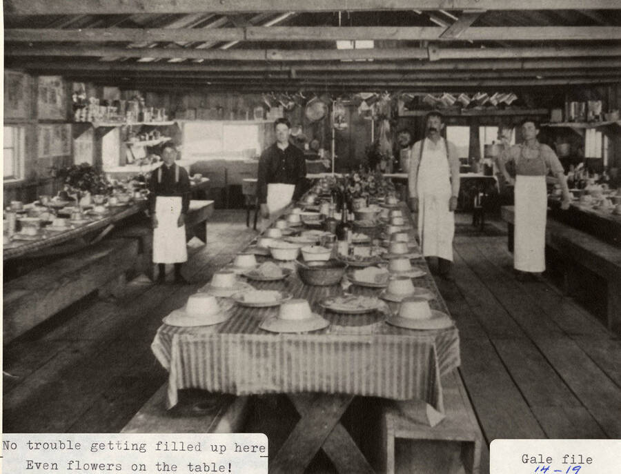 View of the mess hall at a logging camp. Four of the cooks can be seen standing next to fully set tables.
