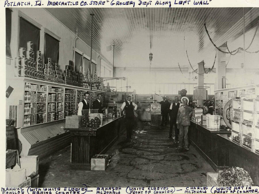 A photograph of the Grocery Department (left wall) of the Potlatch Mercantile Company Store with Manning Fansler, white sleeves behind the counter; Manager McDonald, white sleeves in front of the counter; Charles McDonald, white hat in front of the counter; and others.