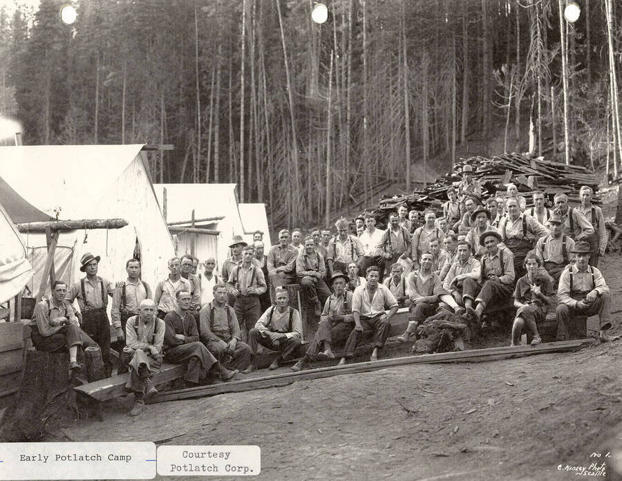 A group of men posing together at one of the early Potlatch camps. Tents can be seen constructed next to them and piles of wood sitting behind them.