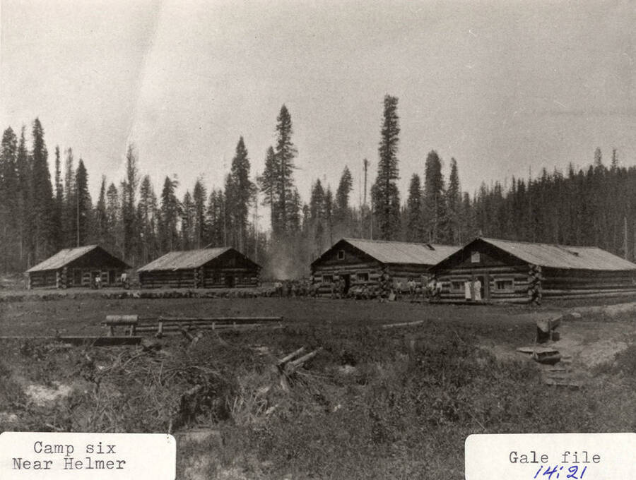 View of Camp 6, which is located near Helmer, Idaho. A group of people can be seen standing in front the log cabins.