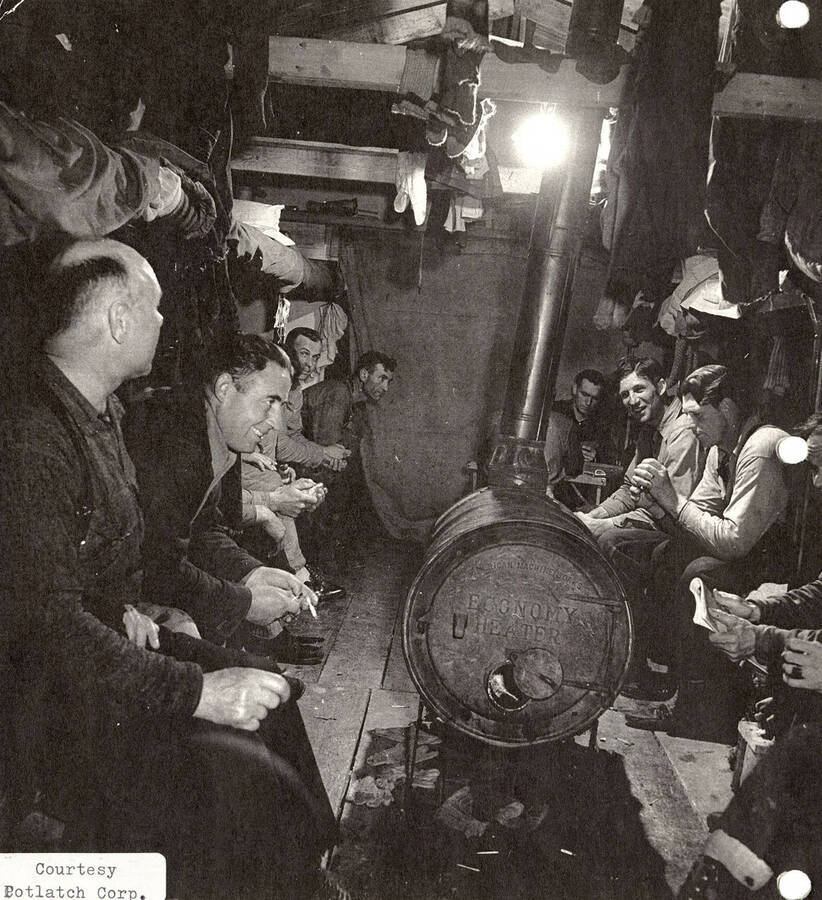 A group of men sitting around a heater in one of the cabins at a PLC camp. Socks and other clothes can be seen hanging from the rafters.