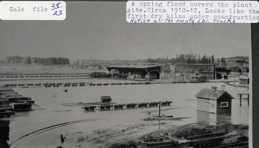 A photograph of the sawmill plant site during a spring flood around 1910-1912. It appears the first dry kilns were under construction and all the lumber trucks are empty.