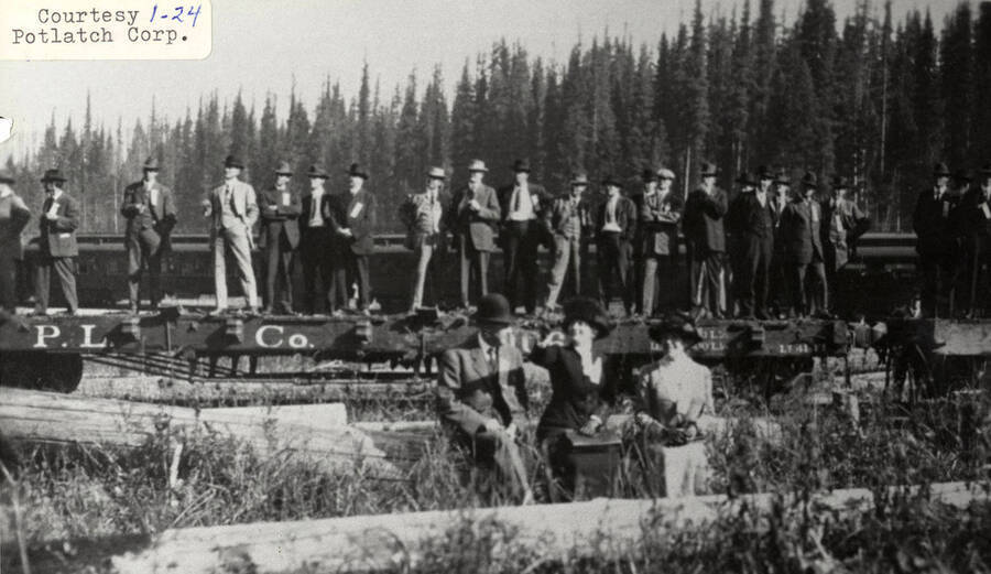 A group of people standing on a few logs at a PLC camp. In the distance a train can be seen approaching on the railroad tracks.