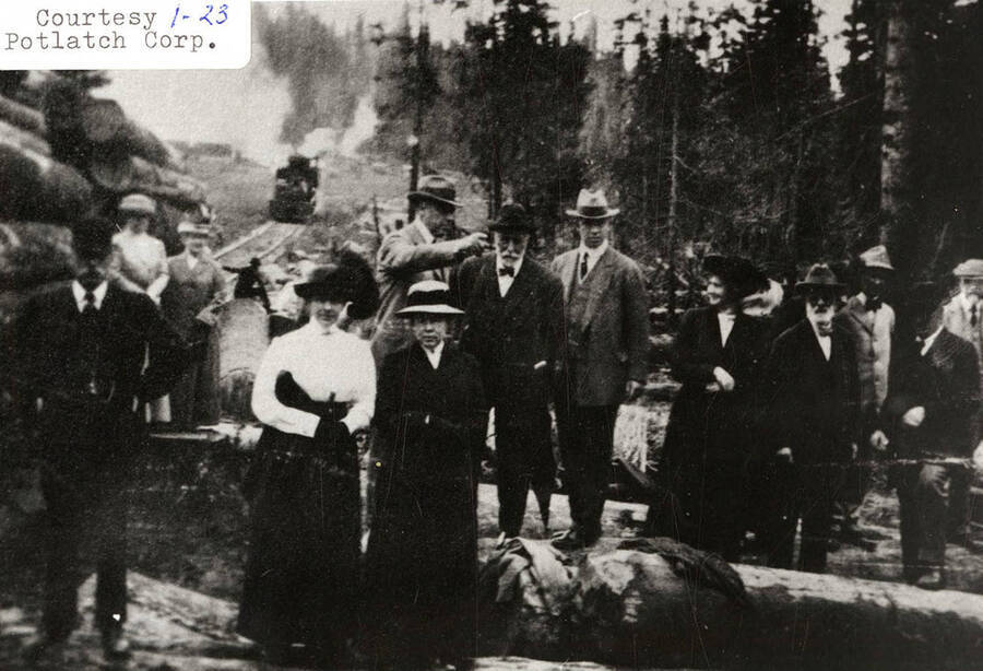 A group of people standing on the railroads in a PLC camp. A few of the people can be seen sitting on logs.