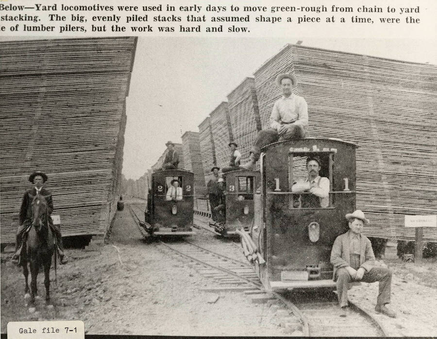 A photograph of employees on the yard locomotives used to move green rough from chain to yard stacking. The piled stacks of lumber were assembled piece at a time with slow hard work.