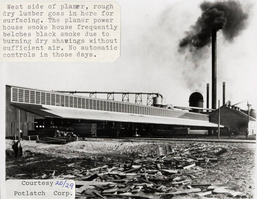 A photograph of the west side of the planer building where rough dry lumber goes for surfacing. The smoke in the background is from the planer power house and was caused by not having enough air to burn the wood shavings properly.