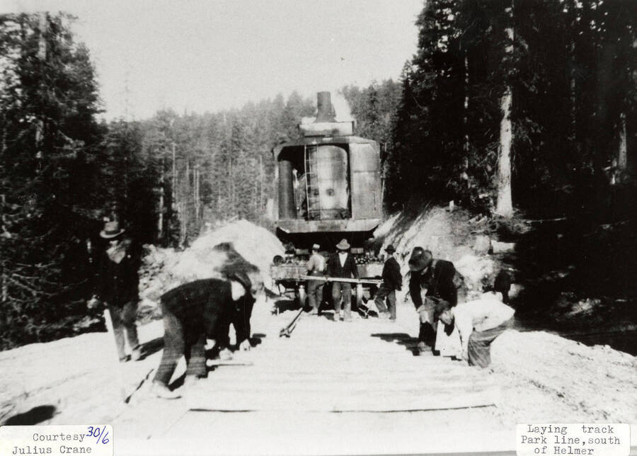 A group of men laying down railroad track at the park line south of Helmer. A railroad car can also be seen on the track.