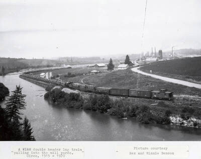 A WI&M doubles header log train pulling logs stacked on flat cars into the mill yards. A river sits to the left of the railroad tracks and houses can be seen to the right.