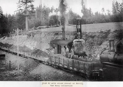 The WI&M No. 2 locomotive with a steam shovel. A few men can be seen sitting on one of the railroad cars and other men can be seen standing around next to dirt piles.
