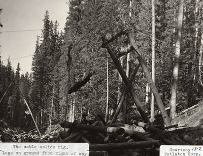 View of the cable splice rig machine. Under the machine, logs lay on the ground in stacks.
