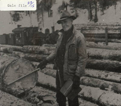A man scaling a log. He can be seen standing among stacks of logs and holding a book in his hand.