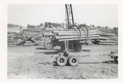 A front view of the LeTourneau log unloading machine with grappling- like hooks in front of several stacks of logs and the town.