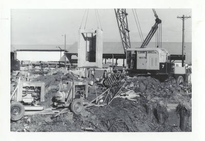 A crane lifting a concrete block missing the side and top with a pendulum like metal piece in the middle.