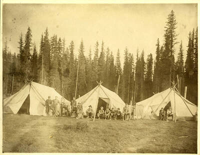 Men posing for the photo outside of three tents made from logs and canvas.