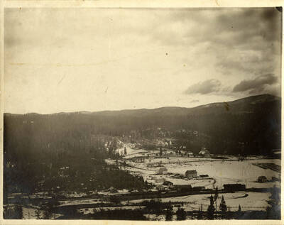 A photograph of tree covered mountains surrounding a snow filled valley with some buildings.