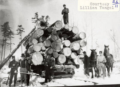 A group of men and a few horses standing around a stack of wood sitting on a railroad track.