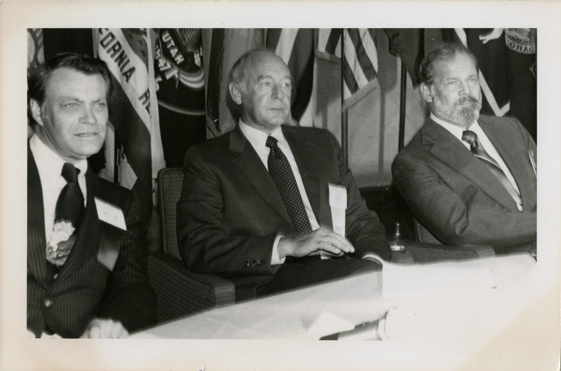 Photo Description: Governor Andrus along with 2 other men sitting together listening to someone talking. There appears to be an arrangement of flags behind them. Text back translation "Governor Cecil D Andrus" Notes read on back "100 Wed Cage B-1 3x3 1/2 Anchorage, Alaska - In a significant session"