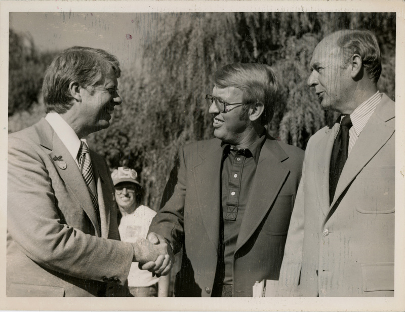 Photo Description: Governor Carter and Attorney General Park are smiling and shaking hands with Governor Andrus (D) right next to them also smiling. Text back translation "Governor Jimmy Carter (D) Attorney General Tony Park (D) Governor Cecil Andrus (D)"