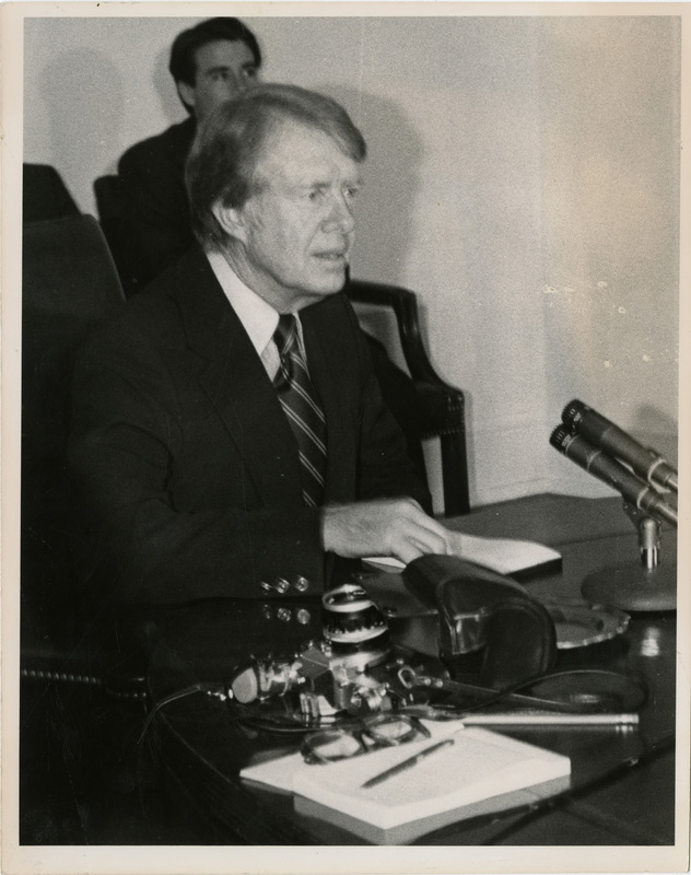 Photo Description:President Jimmy Carter is sitting at a desk in front of the microphone giving a speech. Text back translation "Page e-1 Sunday feature page Washington, D.C. - President Jimmy Carter" Notes read on back "3x7 President Jimmy Carter"