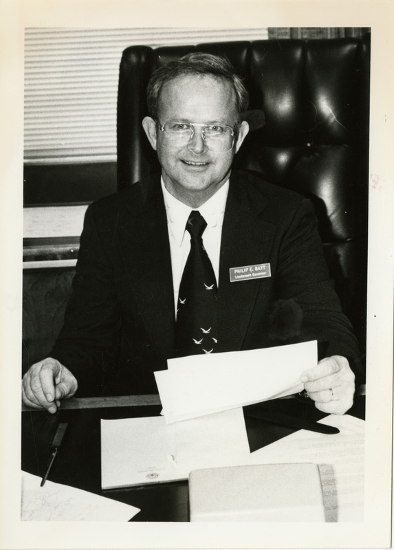 Photo Description: Lt. Governor Philip Batt (R) is reading pictures and sitting at a desk. Notes reads on back "Page A2 100% 2x5 No 2 Official Boise-Lt Gov Phil"