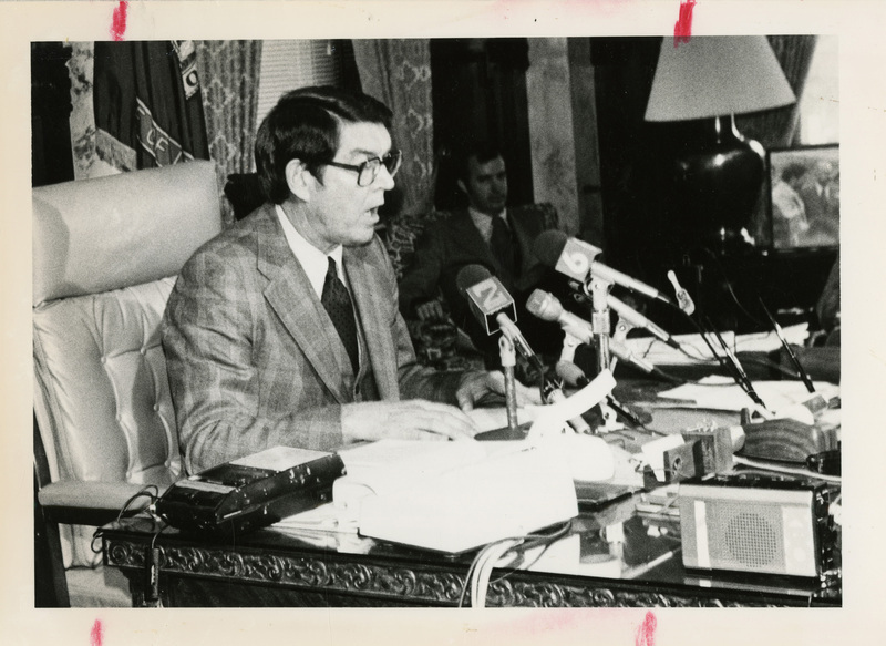 Photo Description: Governor Evans is sitting at a desk with 3 microphones giving a speech. Text back translation "Governor John Evans Angry governor" Notes read on back "2 col as marked Page C-6 2x4 100)