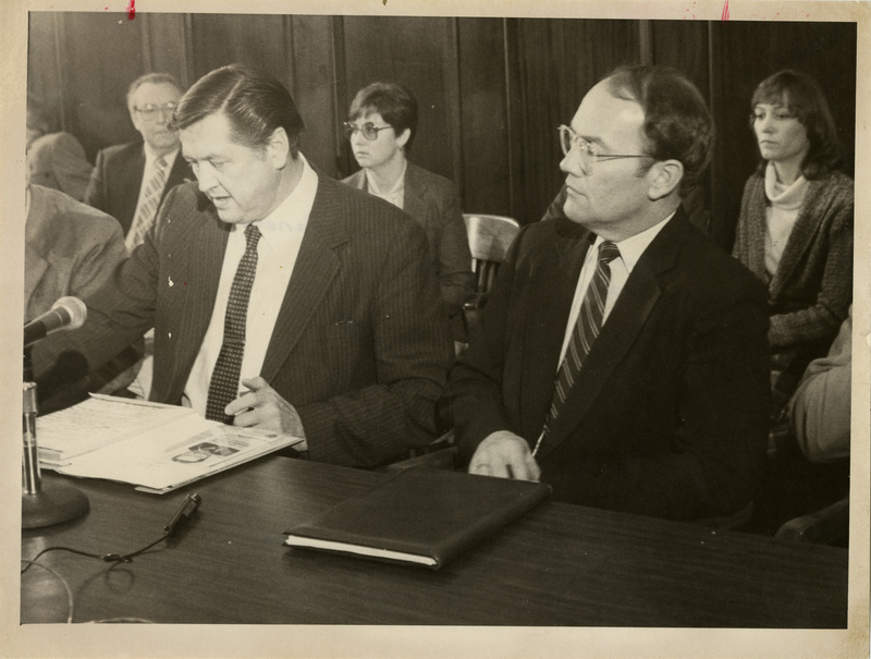 Photo Description: Idaho Representative George Hansen and Senator Larry Craig are sitting at a desk reading a file while answering a question into the microphone. There are a group of people behind them listening. Notes read on back "A-13 Idaho's Congressmen 100% 4x5"