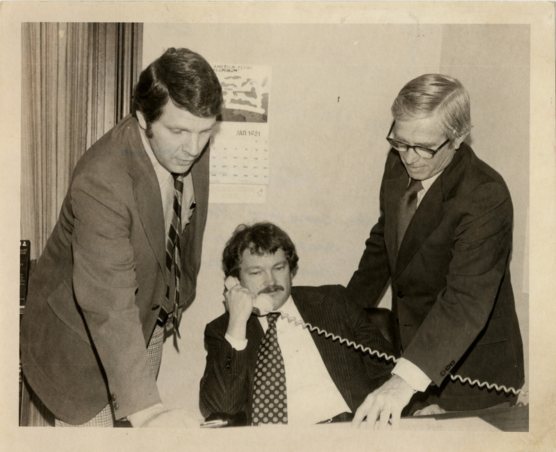 Photo Description: Idaho State Senators Kermit Kiebert (D), Senator Ron Twileger (D), and Senator Mike Mitchell (D) are all crowded around a desk reading some papers. One of them is on leaning back and on the phone. Notes read on back "Page C6 Top Democrat Senators 3x4 1/2 99"