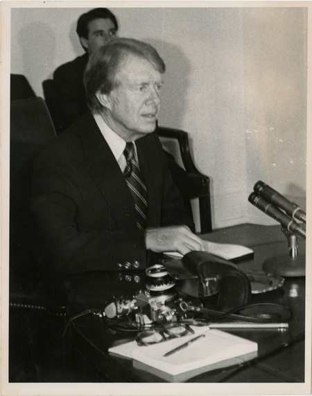 Photo Description:President Jimmy Carter is sitting at a desk in front of the microphone giving a speech. Text back translation "Page e-1 Sunday feature page Washington, D.C. - President Jimmy Carter" Notes read on back "3x7 President Jimmy Carter"