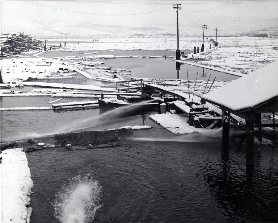In the background snow-covered logs wait to come towards the mill. In the upper left, a pile of logs sits. In the center of the photograph, water sprays on to the pond. The back of the photograph has December 1959 written on it.