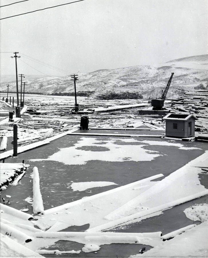 A view of  part of the log pond. The pond in the center of the photograph is frozen and covered in snow, as are the longs and hills surrounding the pond. On the right-hand side of the screen is a crane. On the back of the photograph December 1959 is written.