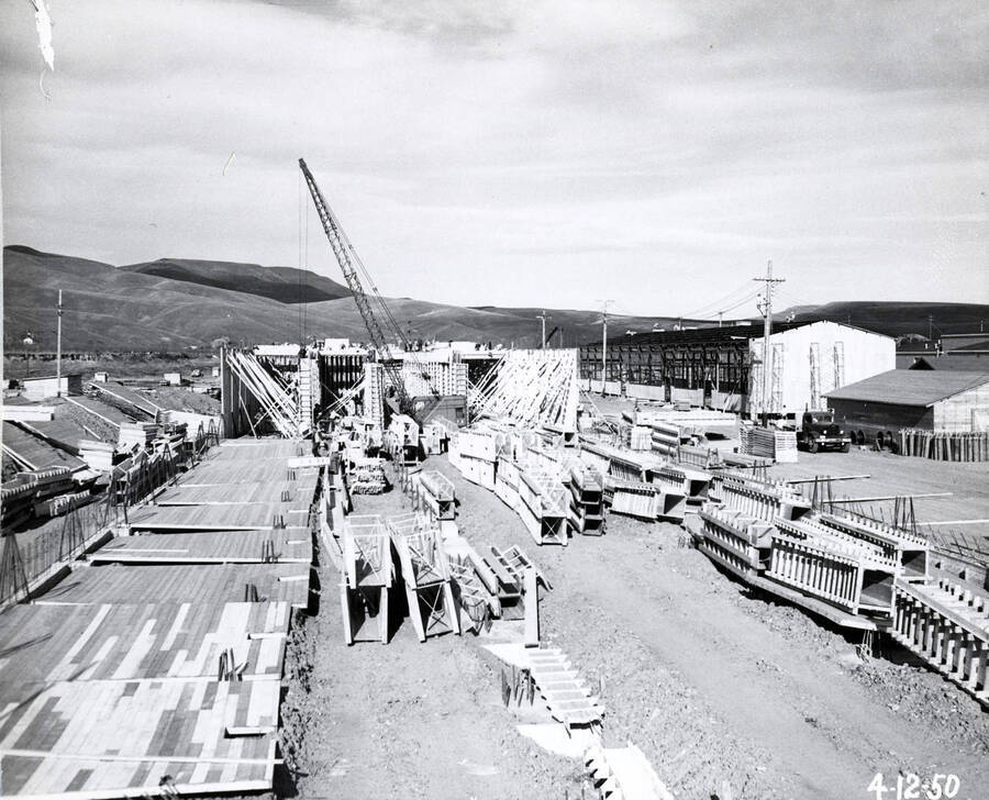 Several of the Clearwater mill's buildings under construction. In the foreground wooden flooring has been laid for one building while in the background framing is happening for another. A crane is assisting with the construction. The date written on the photograph is 4/12/1950.