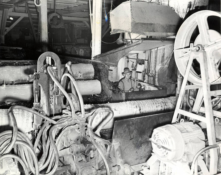 A man sits, operating machinery in the Clearwater mill. In front and to the side of him are debarked logs. The description on the back of the photograph reads 'Headrig #1, night sawyer B.W. Sebring.'