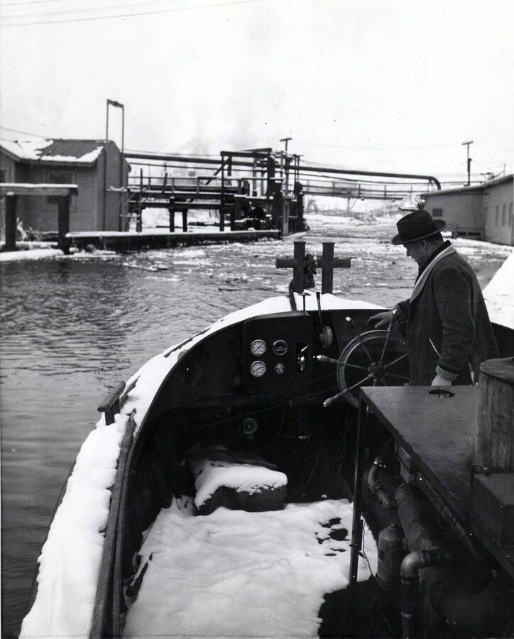 A bundled up mill worker drives a snow-covered boat in the log pond. The back of the photograph lists the date as December 1959.