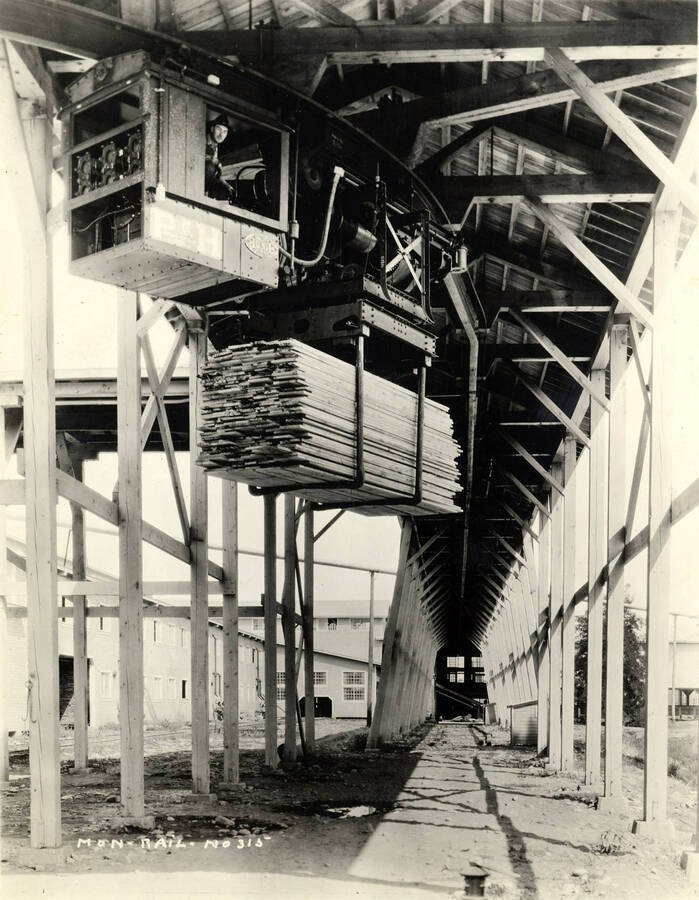 A driver looks out the window of the engine car of a monorail. The piece of equipment in front of him holds a stack of planks being moved to a different location within the mill grounds. Written on the photograph is 'Mon rail, No. 315'.
