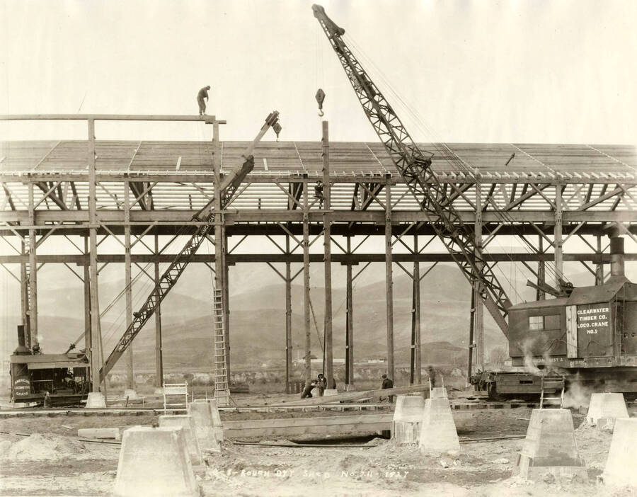 Construction of the rough-dry shed at the Lewiston Mill. Two cranes are being used while three men work on the roof. Four men stand on the ground in the middle of the picture working. Written on the picture is 3-8 Rough dry shed, No. 211 1927.