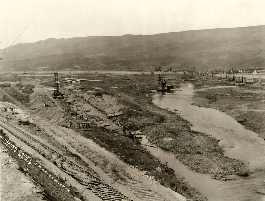 Construction of unloading docks in what would be the log pond. Men work on laying the structures that would eventually carry railcars full of logs to dump into the log pond. At the end of the line sits a crane-like device. In the foreground of the picture sit two railroad tracks. Written on the photograph is 'Unloading docks, CT CO Lewiston, 3/16/1927 No. 218.'