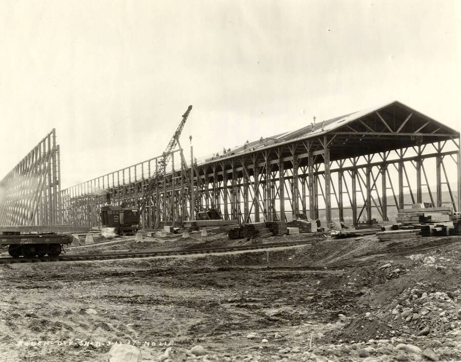 Construction of the Rough-dry shed at the Lewiston mill. A crane is assisting with building the roof on the frame constructed building. Piles of lumber sit around the building as well. Written on the photograph is 'Rough dry shed, 3/31/1927, No. 225.'