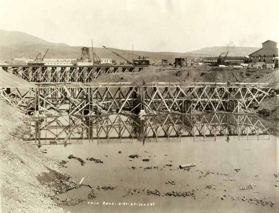 A man pushes a wheeled cart over a wooden bridge. In the background, a crane is helping with another area of construction. Written on the photograph is 'Tail Race, 3/30/1927 No. 237.'