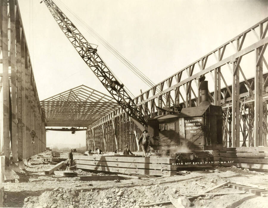 A view inside the rough-dry shed. A Crane is working pulling lumber to the top for the roof. A man stands beside the crane. Written on the photograph is 'Rough dry shed, 3/28/1927, No. 235.'