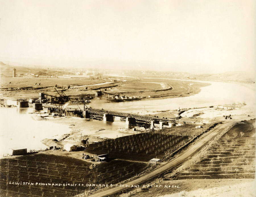 View of the plant facing Lewiston. The plant can be seen in the background. In the foreground is the Lewiston Dam under construction as well as rows of trees. Written on the photograph is 'Lewiston Power and Light Company. Dam and CT CO Plant. 2/24/1927 No. 206.'