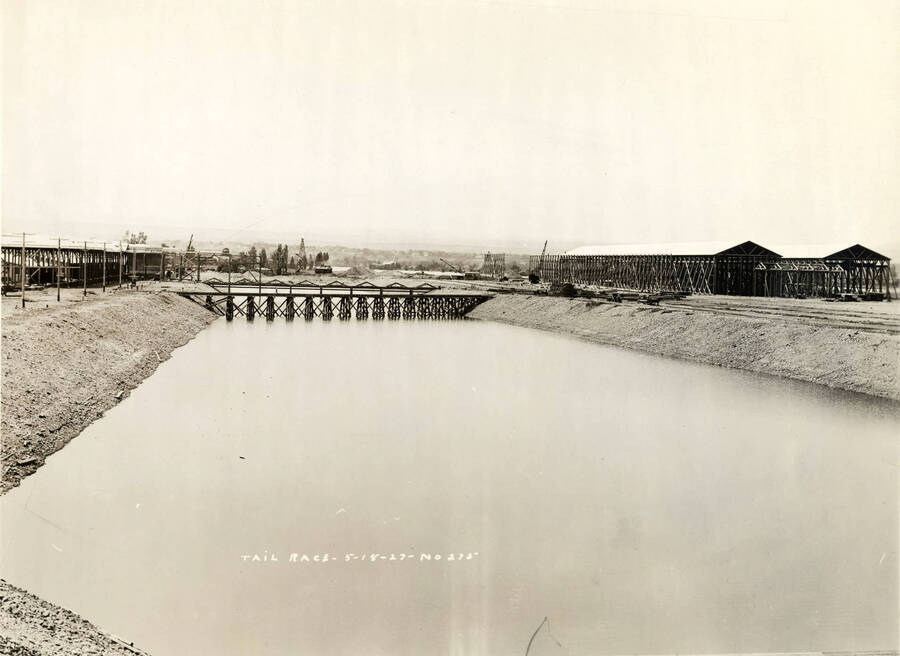 View of the bridge across the part of the Clearwater river that runs near the plant. On the right hand side are two frame constructed buildings. On the left hand side are partially constructed buildings. Written on the photograph is 'Tail Race, 5/18/1927, No. 275.'