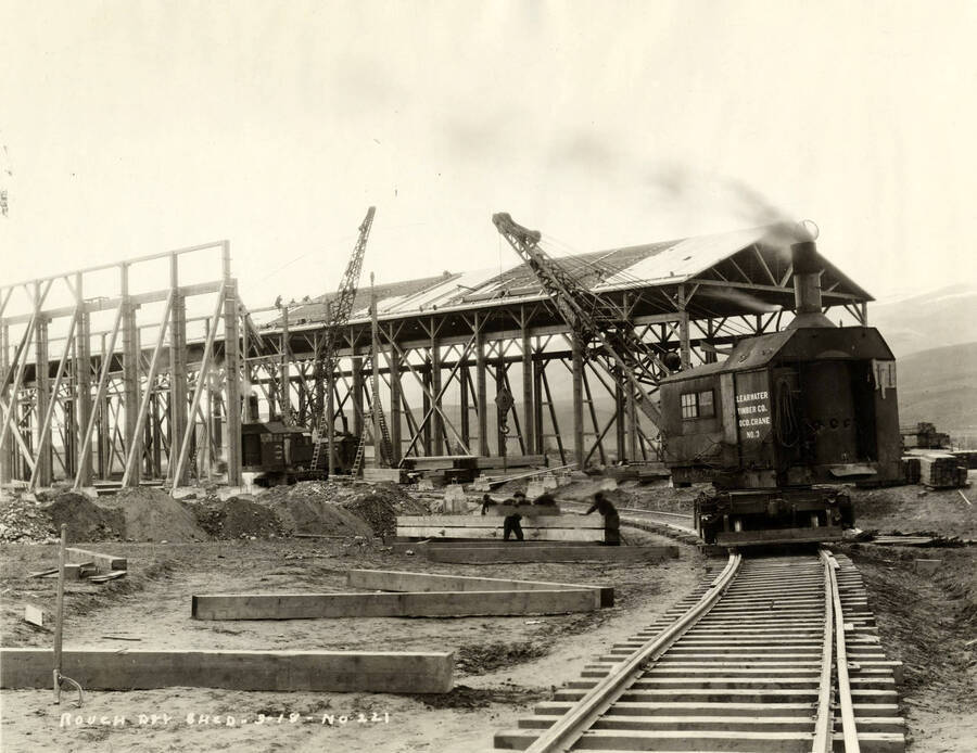 Construction of the rough-dry shed in progress. One crane in the background works on the building lifting roofing materials, while another in the foreground lifts lumber for the other building. Written on the photograph is 'Rough dry shed, 3/18/1927, No. 221.'