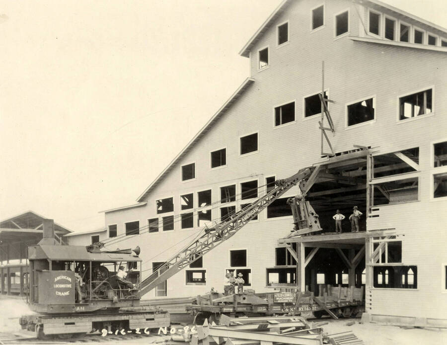 Men use a crane to hoist machinery up on to the second floor on of one of the buildings of the Lewiston Mill. In front of the train are rail cars where three men stand. Written on the crane is 'American Locomotive Crane.' Written on the photograph is '8/16/1926 No. 96'.