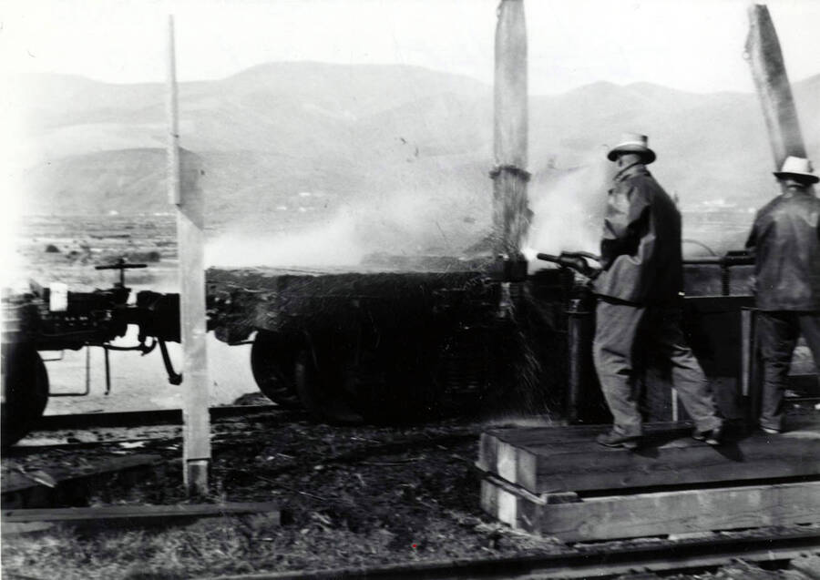 Men work to spray bark off of flatcars that brought in logs. Date of the back of the picture says it was taken in April.