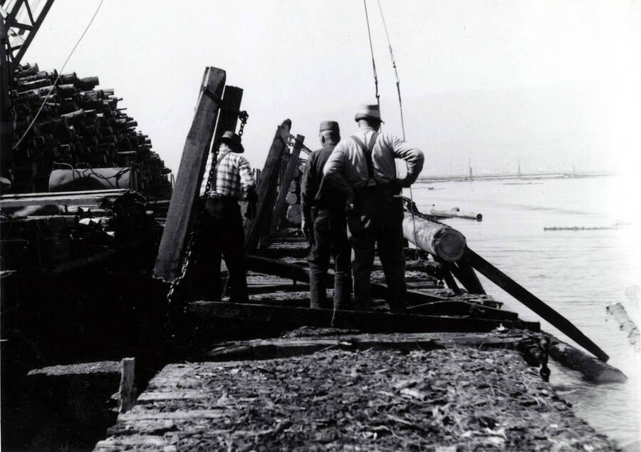 Men work to attach crane lines to a log. To the right of the men is the log pond.